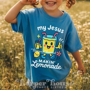 Op de afbeelding: Een blauw t-shirt met de tekst "my Jesus makin' lemonade" en een cartoon limonade afbeelding. Het shirt wordt gedragen door een kind dat een madeliefje vasthoudt. De woorden "Original Designer" staan bovenaan.