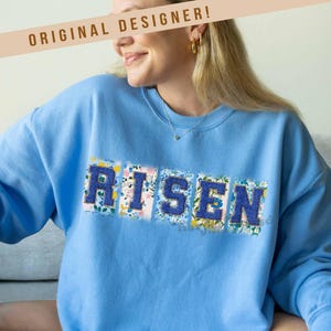 May include: A light blue sweatshirt with the word "RISEN" spelled out in floral patterned letters. The sweatshirt is made by Harper House Three Sixteen.