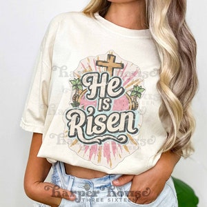 May include: A white t-shirt with a vintage-style graphic design featuring a cross, palm trees, and the text "He is Risen".