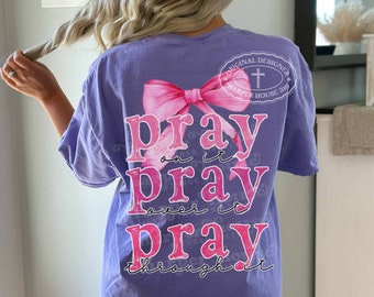 Könnte beinhalten: Ein lilafarbenes T-Shirt mit einer rosa Schleife und den Worten "pray on it, pray over it, pray through it" in Pink. Das Shirt hat auch ein Logo mit der Aufschrift "Original Designer Harper House 316".