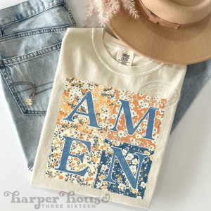 May include: A beige t-shirt with a floral pattern design that spells out the word "AMEN" in blue letters. The shirt is photographed on a flat surface with a pair of blue jeans and a straw hat.