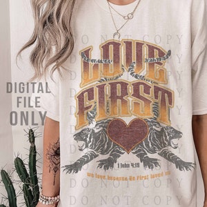May include: A white t-shirt with a graphic design of two tigers with a heart in the middle. The text "Love First" is written in a vintage font above the tigers. The text "1 John 4:19" is written below the tigers. The text "We love because He first loved us" is written below the scripture reference. The text "Do Not Copy" is repeated in a diagonal pattern around the graphic design.