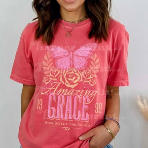Pu&ograve; includere: Una maglietta rosa corallo con una stampa grafica di una farfalla, rose e il testo "Amazing Grace 1999 How Sweet The Sound".