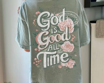 May include: A sage green short-sleeve t-shirt featuring a back graphic with the text God is Good all the Time in white lettering. The design includes illustrations of pink roses, green leaves, and small red berries, with a cross symbol.