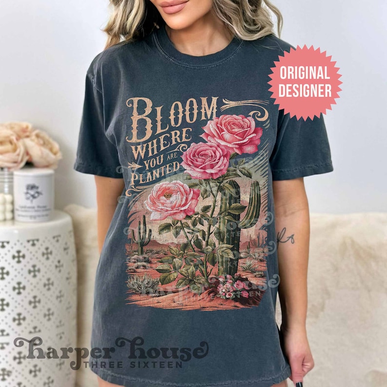 May include: A dark gray t-shirt with a vintage-style graphic of roses and a cactus. The text on the shirt reads "Bloom Where You Are Planted".