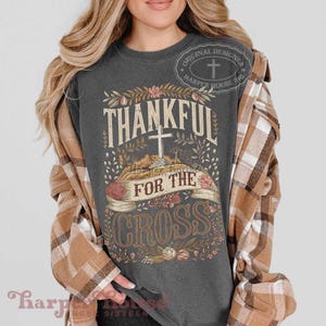 Op de afbeelding: Een antracietgrijs t-shirt met korte mouwen en een grafische print met de tekst Thankful For The Cross, een centrale kruisillustratie, bloemaccenten en een banner. Een bruin-wit geruit flanellen overhemd wordt eroverheen gedragen.