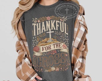 Puede incluir: Una camiseta gris carbón de manga corta con un estampado gráfico que incluye el texto Thankful For The Cross, una ilustración de una cruz central, detalles florales y una cinta. Se lleva una camisa de franela a cuadros marrón y blanca.