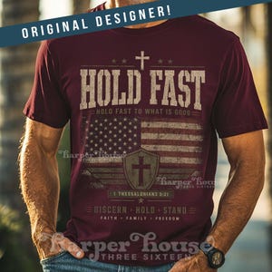 May include: Maroon t-shirt with a distressed American flag design and the words "HOLD FAST." The shirt includes a cross, stars, and the text "HOLD FAST TO WHAT IS GOOD." Additional text includes "1 Thessalonians 5:21" and "DISCERN HOLD STAND."