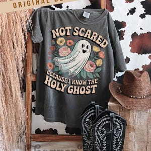 May include: Dark gray t-shirt with the text "NOT SCARED BECAUSE I KNOW THE HOLY GHOST" and a cartoon ghost illustration surrounded by flowers. The shirt is displayed with cowboy boots and a hat.