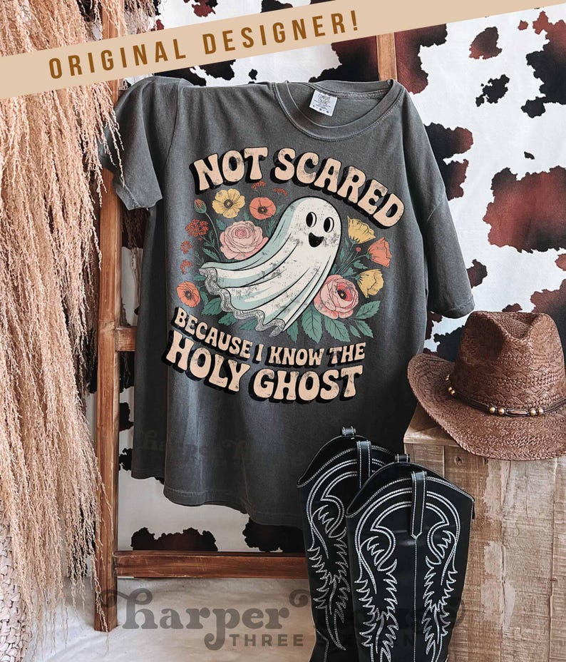 May include: Dark gray t-shirt with the text "NOT SCARED BECAUSE I KNOW THE HOLY GHOST" and a cartoon ghost illustration surrounded by flowers. The shirt is displayed with cowboy boots and a hat.