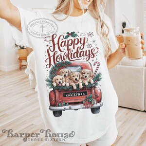 Op de afbeelding: Wit T-shirt met een rode vrachtwagen die vier golden retriever-puppies vervoert. De vrachtwagen is versierd met een kerstkrans en de tekst "Happy Howlidays" en "Christmas".