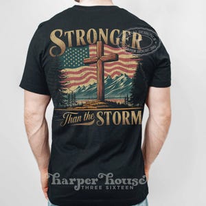 May include: A black t-shirt with a vintage-style graphic of a cross in front of an American flag. The text "Stronger Than the Storm" is printed below the graphic. The shirt has a distressed look and is perfect for anyone who wants to show their faith and patriotism.