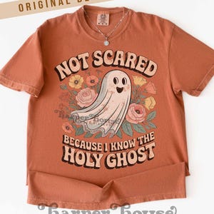 May include: Terracotta-colored t-shirt with the text "NOT SCARED BECAUSE I KNOW THE HOLY GHOST" and a graphic of a friendly ghost surrounded by flowers. The shirt has short sleeves and a round neckline. A silver necklace is visible.