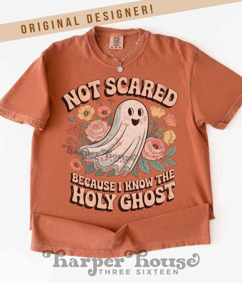 May include: Terracotta-colored t-shirt with the text "NOT SCARED BECAUSE I KNOW THE HOLY GHOST" and a graphic of a friendly ghost surrounded by flowers. The shirt has short sleeves and a round neckline. A silver necklace is visible.