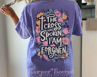 May include: A light purple short-sleeve t-shirt featuring a large back graphic of colorful flowers surrounding the text: The Cross Has Spoken I Am Forgiven Ephesians 1:7. The design uses white and dark blue lettering with pink and orange floral accents.