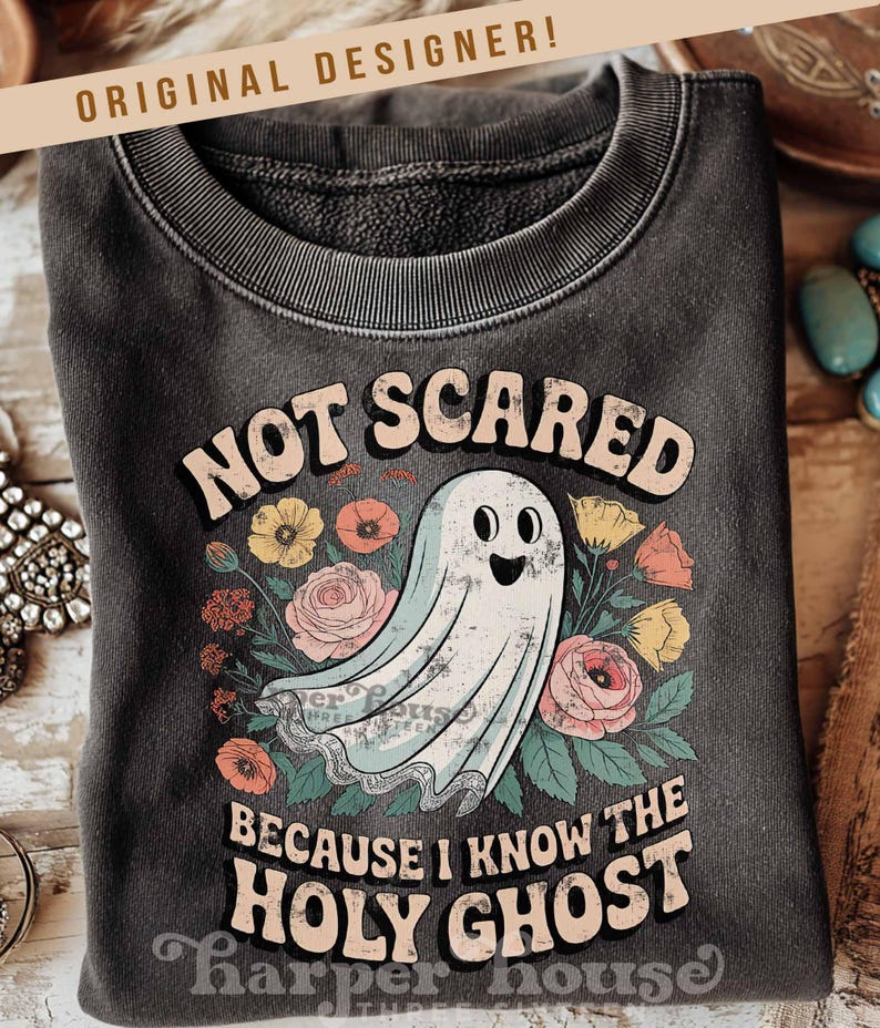 May include: Dark gray sweatshirt with the text "NOT SCARED BECAUSE I KNOW THE HOLY GHOST" and a cartoon ghost surrounded by flowers. The words "ORIGINAL DESIGNER!" are at the top. The sweatshirt is folded.