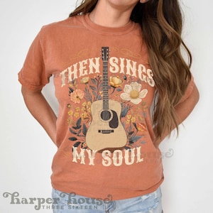 May include: A burnt orange t-shirt with a floral design featuring a guitar and the text "Then Sings My Soul".