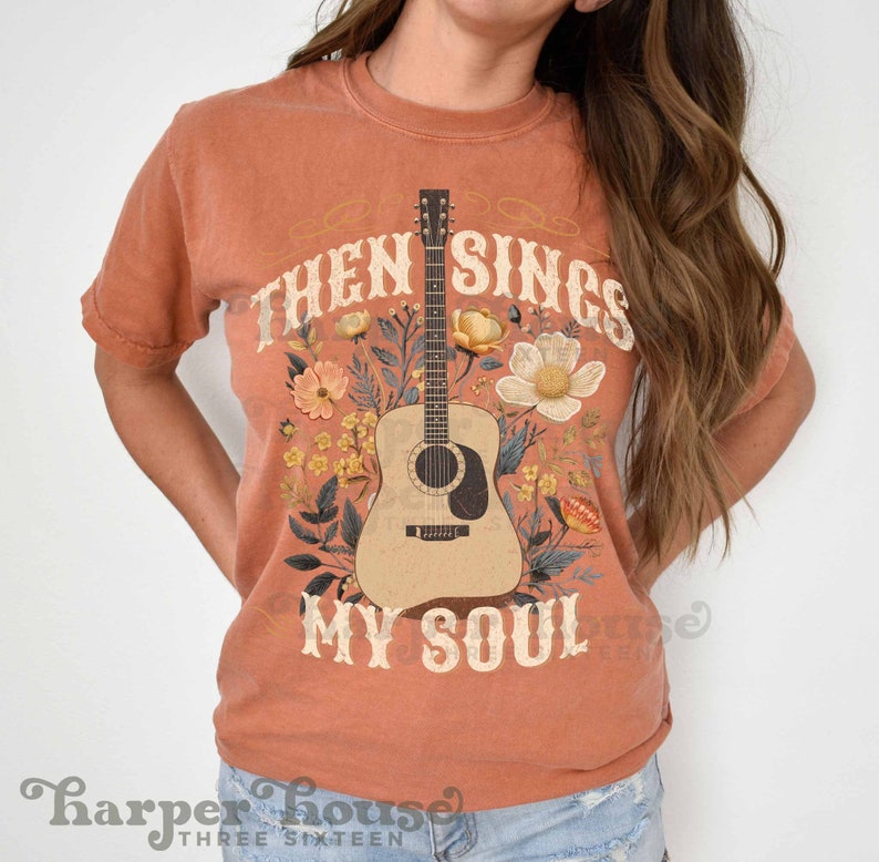May include: A burnt orange t-shirt with a floral design featuring a guitar and the text "Then Sings My Soul".
