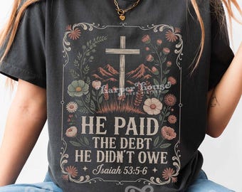 May include: A charcoal gray t-shirt featuring a graphic print of a wooden cross, mountains, and floral elements. The text reads: HE PAID THE DEBT HE DIDN'T OWE Isaiah 53:5-6. The design is framed by decorative scrollwork and botanical illustrations.