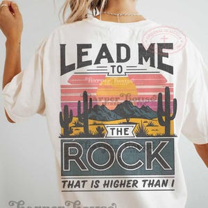 May include: White t-shirt with a vintage-style graphic that says "Lead me to the rock that is higher than I". The graphic features a sunset, mountains, and cacti.