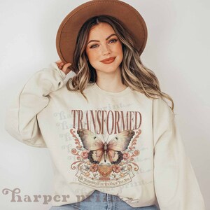 May include: A cream-colored sweatshirt with a graphic print of a butterfly with pink flowers and the word "TRANSFORMED" in red. The text below the butterfly reads "Be transformed by the renewing of your mind."