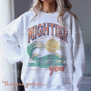 May include: A white crewneck sweatshirt featuring a graphic print with the text MIGHTIER THAN THE WAVES OF THE SEA IS HIS LOVE FOR YOU Psalm 93:4. The design includes a large green ocean wave, a yellow sun, and distressed lettering.