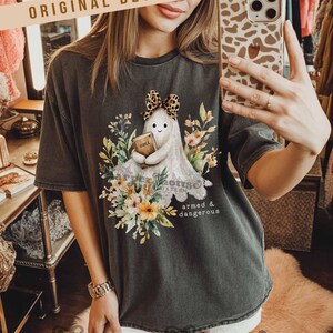 May include: Dark gray t-shirt featuring a cartoon ghost holding a book labeled "Bible" and wearing a leopard print bow. The ghost is surrounded by floral elements and the text "armed & dangerous". The shirt has the text "ORIGINAL DESIGNER!" at the top.