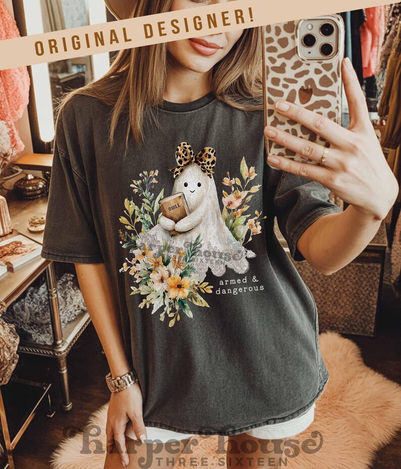 May include: Dark gray t-shirt featuring a cartoon ghost holding a book labeled "Bible" and wearing a leopard print bow. The ghost is surrounded by floral elements and the text "armed & dangerous". The shirt has the text "ORIGINAL DESIGNER!" at the top.