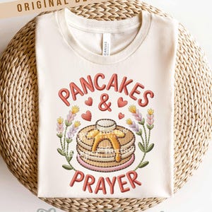 May include: Cream-colored t-shirt with the embroidered text "PANCAKES & PRAYER". The design features a stack of pancakes with butter, hearts, and floral accents. The words "ORIGINAL DESIGNER!" are at the top.