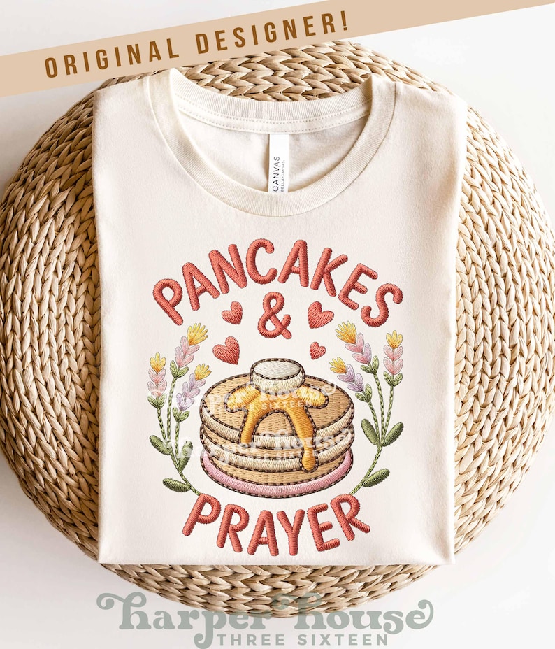 May include: Cream-colored t-shirt with the embroidered text "PANCAKES & PRAYER". The design features a stack of pancakes with butter, hearts, and floral accents. The words "ORIGINAL DESIGNER!" are at the top.