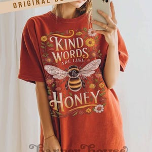 Peut inclure: T-shirt rouille avec un motif d'abeille et les mots "Kind Words Are Like Honey". Le graphisme comprend des &eacute;l&eacute;ments floraux. Le t-shirt a des manches courtes et une coupe d&eacute;contract&eacute;e.