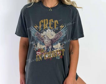 May include: A charcoal gray short-sleeve t-shirt featuring a graphic of a bald eagle with spread wings, gold lightning bolts, and red roses. The text "FREE IN CHRIST" is printed in gold lettering above and below the eagle illustration.