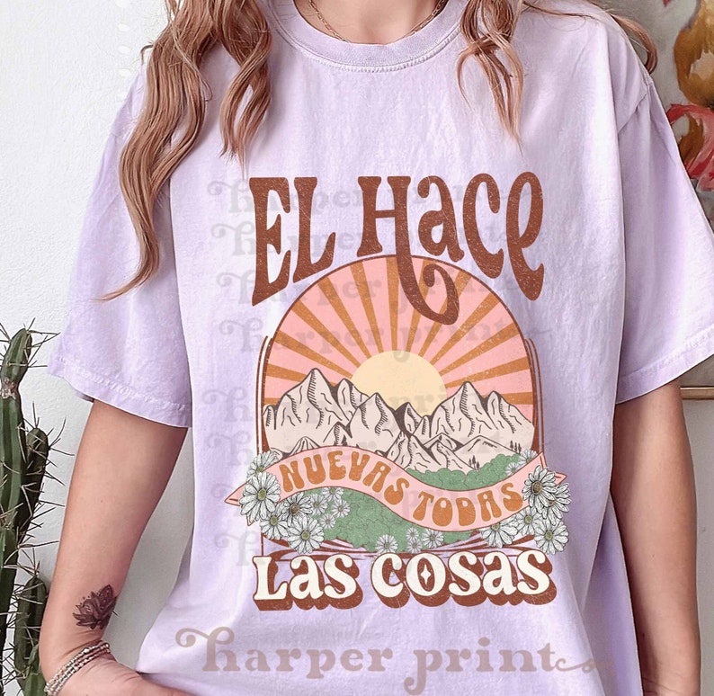 May include: A lavender t-shirt with a graphic print of a sunset over mountains with the text "El Hace Nuevas Todas Las Cosas" in brown lettering.