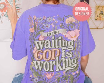 Puede incluir: Una camiseta de color morado claro con un gráfico en la espalda de flores silvestres coloridas que rodean el texto "in the waiting GOD is working". El diseño utiliza una mezcla de letras cursivas y negritas en tonos rosa, blanco y verde.