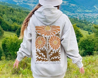 Puede incluir: Una sudadera con capucha gris claro que presenta un gráfico grande en la espalda con tres flores y hojas estilizadas de color naranja y marrón. Debajo del diseño floral, el texto dice: Let us not become weary in doing good, for at the proper time we will reap a harvest if we do not give up. Galatians 6:9.