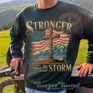 May include: Green sweatshirt with a distressed American flag and a wooden cross. The text "Stronger Than the Storm" is printed below the flag. The text "Original Designer" is printed in a circle above the flag.