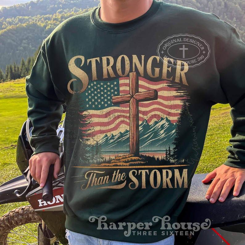 May include: Green sweatshirt with a distressed American flag and a wooden cross. The text "Stronger Than the Storm" is printed below the flag. The text "Original Designer" is printed in a circle above the flag.