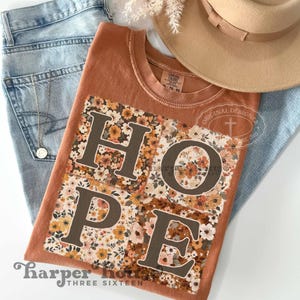 May include: A brown t-shirt with a patchwork design featuring the word "HOPE" in brown letters. The patchwork is made up of floral patterns in shades of orange, yellow, and brown.