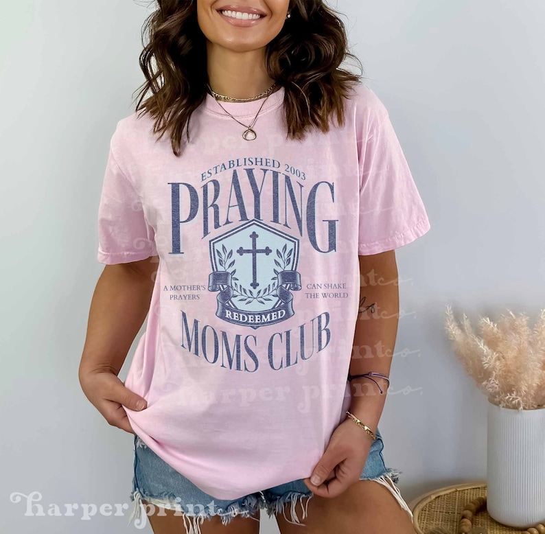 Op de afbeelding: Roze T-shirt met een distressed look met een blauwe en witte afbeelding met de tekst "Established 2003 Praying Redeemed Moms Club". De afbeelding bevat een kruis en een krans.