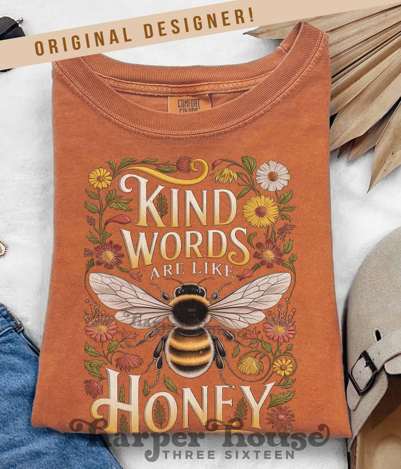 Peut inclure: Un t-shirt orange avec le texte "Kind Words Are Like Honey" et une illustration d&eacute;taill&eacute;e d'une abeille. Le motif est entour&eacute; d'&eacute;l&eacute;ments floraux. Le t-shirt a un col rond et des manches courtes.