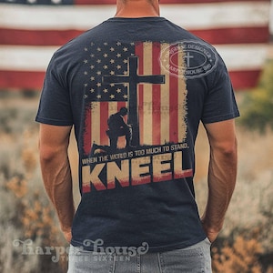 Pode incluir: Uma camiseta azul marinho com um design de bandeira americana desbotada que apresenta uma cruz e um soldado ajoelhado. O texto "WHEN THE WORLD IS TOO MUCH TO STAND, KNEEL" &eacute; impresso abaixo da imagem. A camiseta tamb&eacute;m tem um pequeno logotipo que diz "ORIGINAL DESIGNER HARPE HOUSE 316".