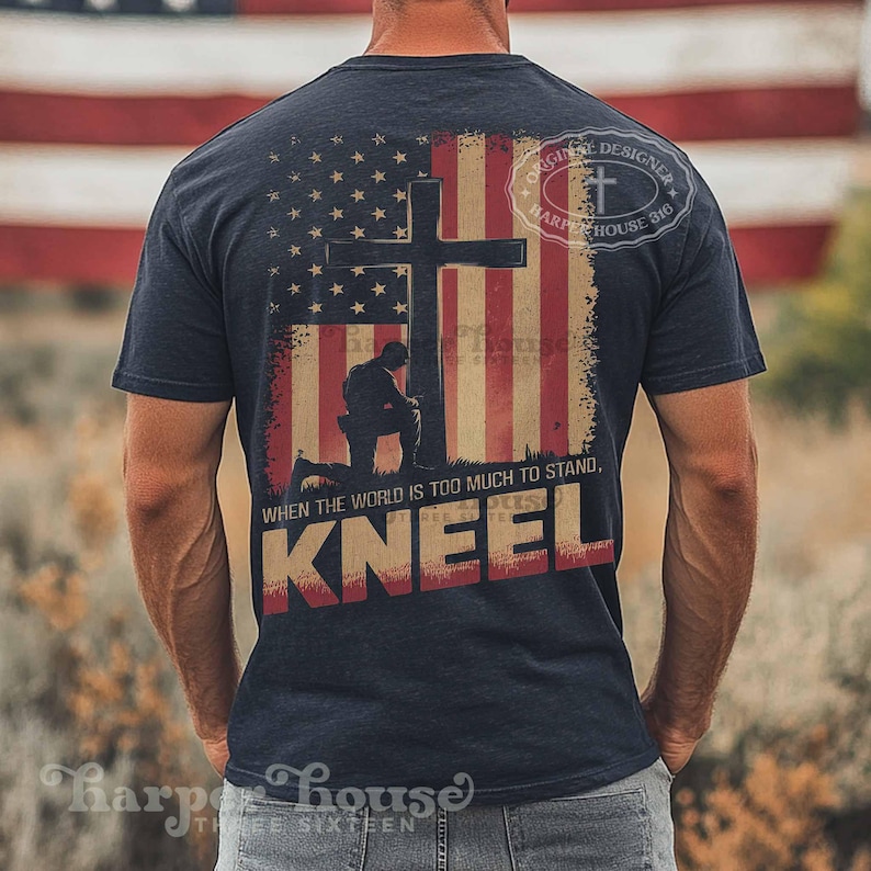 Pode incluir: Uma camiseta azul marinho com um design de bandeira americana desbotada que apresenta uma cruz e um soldado ajoelhado. O texto "WHEN THE WORLD IS TOO MUCH TO STAND, KNEEL" &eacute; impresso abaixo da imagem. A camiseta tamb&eacute;m tem um pequeno logotipo que diz "ORIGINAL DESIGNER HARPE HOUSE 316".