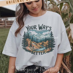 May include: White t-shirt with a nature-themed graphic. The design features a mountain range, trees, and a river, with the text "YOUR WAY'S BETTER". The shirt also has the text "ORIGINAL DESIGNER!" at the top.