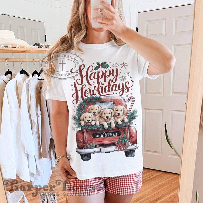 Op de afbeelding: Wit T-shirt met een rode vrachtwagen die vier golden retriever-puppies vervoert. De vrachtwagen heeft de tekst "Christmas" op de achterkant. De tekst op het T-shirt luidt "Happy Howlidays".