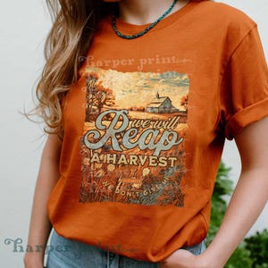 Op de afbeelding: Een roestoranje t-shirt met korte mouwen en een grafische print van een boerderijlandschap met een schuur, korenvelden en de tekst "We will Reap A HARVEST" en "WE DON'T GIVE UP". Het shirt heeft een ronde hals en een relaxte pasvorm.