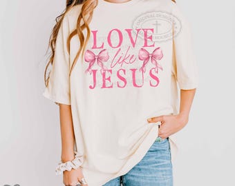 May include: An off-white short-sleeve t-shirt featuring the pink text LOVE like JESUS and two pink bows. The design includes a circular watermark with a cross symbol. The shirt is worn with blue denim jeans and a white wrist scrunchie.