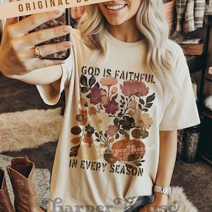 May include: Cream-colored t-shirt with a floral and pumpkin graphic. The text on the shirt reads "GOD IS FAITHFUL IN EVERY SEASON." The design includes flowers in shades of pink, burgundy, and cream. The shirt has short sleeves.