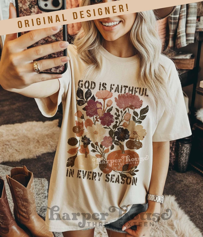 May include: Cream-colored t-shirt with a floral and pumpkin graphic. The text on the shirt reads "GOD IS FAITHFUL IN EVERY SEASON." The design includes flowers in shades of pink, burgundy, and cream. The shirt has short sleeves.