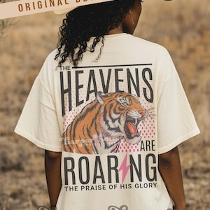 May include: Cream-colored t-shirt with a tiger graphic and the words "THE HEAVENS ARE ROARING" in black. A pink lightning bolt is below the word "ROARING". The text "THE PRAISE OF HIS GLORY" is below the word "ROARING".