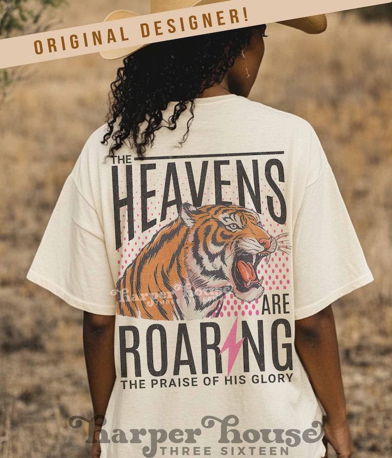 May include: Cream-colored t-shirt with a tiger graphic and the words "THE HEAVENS ARE ROARING" in black. A pink lightning bolt is below the word "ROARING". The text "THE PRAISE OF HIS GLORY" is below the word "ROARING".